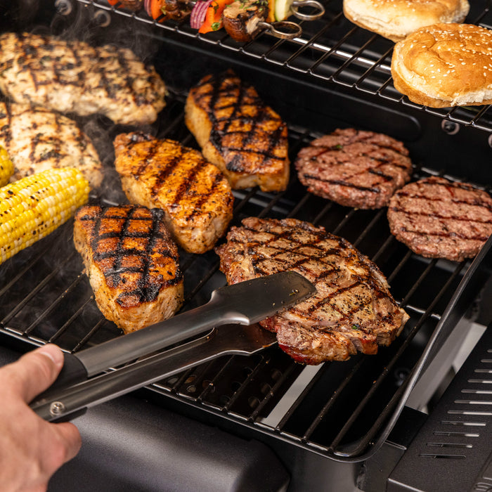 Black tongs flipping grilled steak on a multi-level grill with burgers, corn, and vegetables.