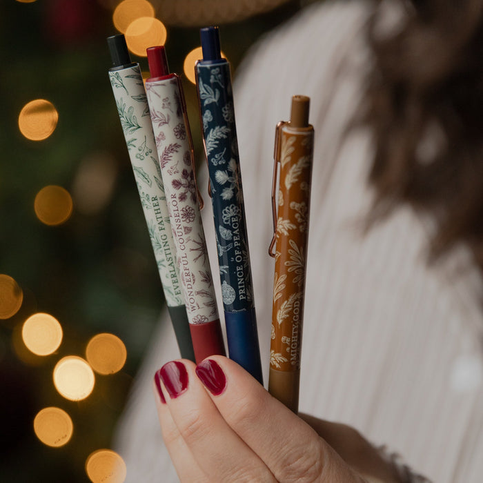 A close-up of a hand holding four decorative ballpoint pens with floral patterns, set against a warm, blurred Christmas tree background with glowing lights.