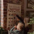 A woman and child hanging a fabric Advent calendar on a brick wall, with the woman pointing to one of the numbered pockets. The calendar has multiple small ornaments inside and the text 'Come Thou And Let Us Expect Thy Jesus' at the top.