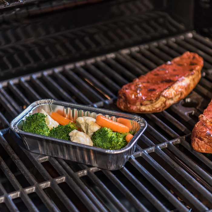 Grilled steak with foil tray of broccoli, cauliflower, and carrots on barbecue grill.
