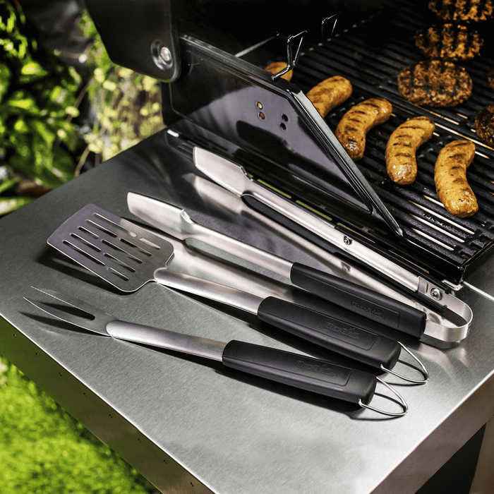 "Set of black-handled stainless steel grilling tools on a grill with sausages."