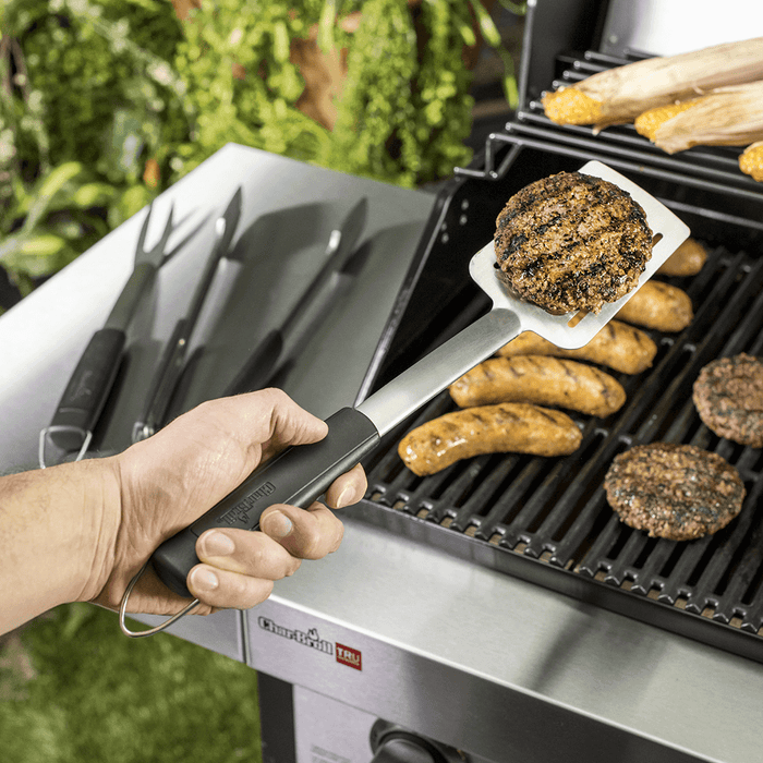 Char-Broil black spatula holding burger patty on grill with sausages and corn.