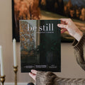 Person holding 'be still' magazine issue 38 in a room with candles and a painting.