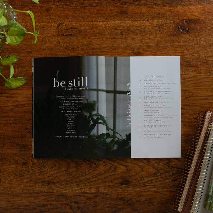 Be Still Issue 40 Magazine opened to the index page on a wooden table near some greenery