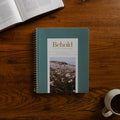 Behold Volume 3 Spiral cover on a wooden table near a coffee cup and an open bible