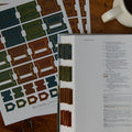 A Bible study or prayer journal shown open on a wooden table, with printed pages visible and matching printed sheet cutouts laid beside it. The book has colored page headers in blue, green, brown, and gold, and the visible title reads 'The Daily Grace Journal.
