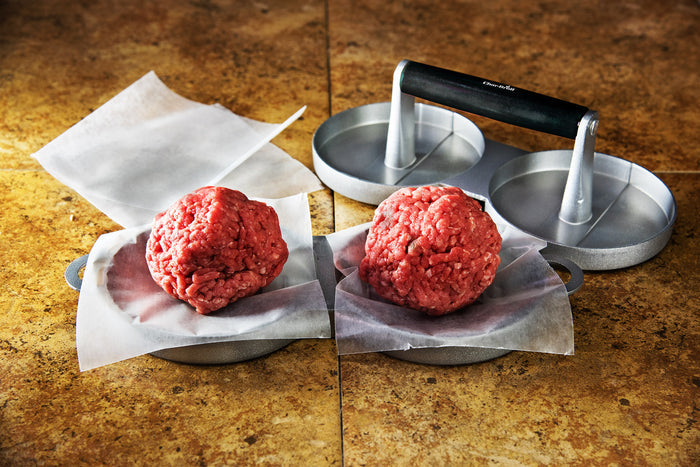 Silver meatball press with black handle, forming red meatballs on parchment paper.