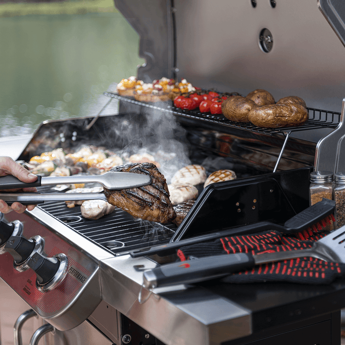 Red and black performance grill with tongs lifting steak, vegetables, and potatoes cooking.