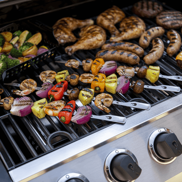 Stainless steel gas grill with chicken, sausages, and vegetable skewers cooking.