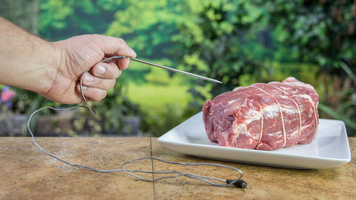 Gray meat thermometer probe inserted into raw meat on a plate.