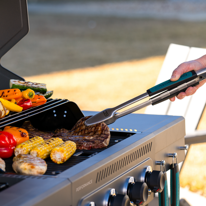 Green and black tongs on a grill with steak and vegetables.