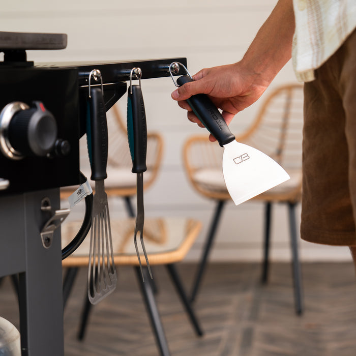 Black OXO Good Grips spatula with ergonomic handle, hanging on a grill accessory rack.
