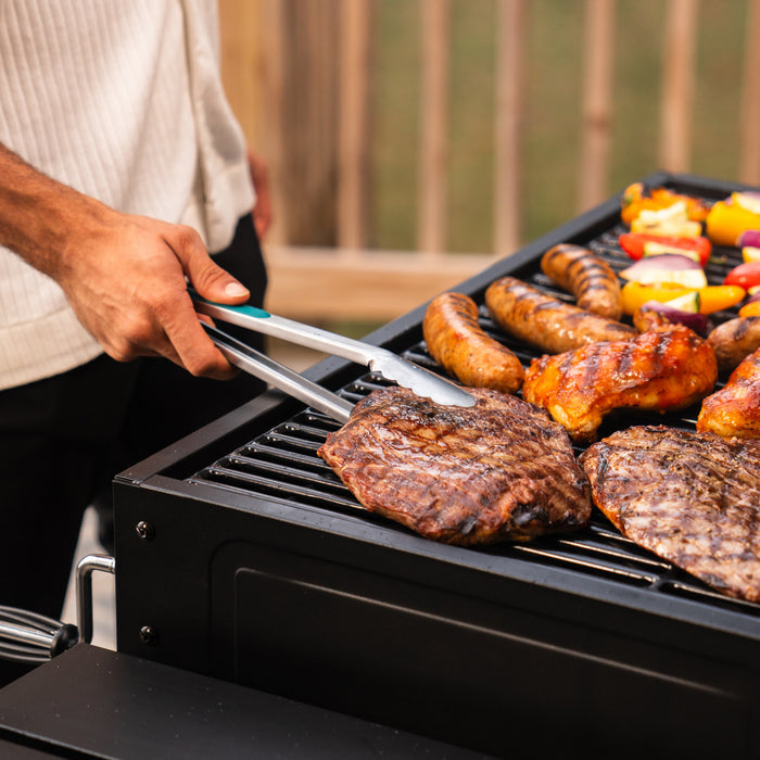 Black grill with tongs flipping steak and sausages, colorful vegetables on side.