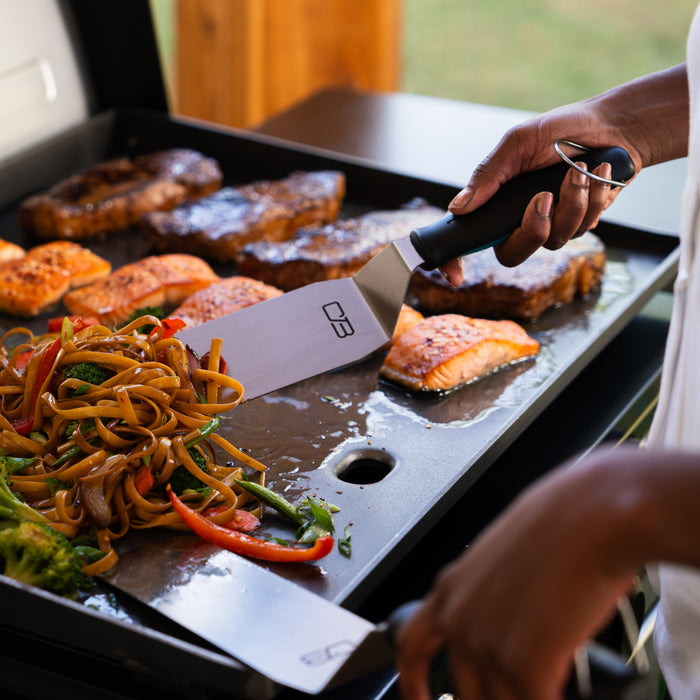 "Black-handled spatula with silver blade on a griddle with salmon and vegetables."