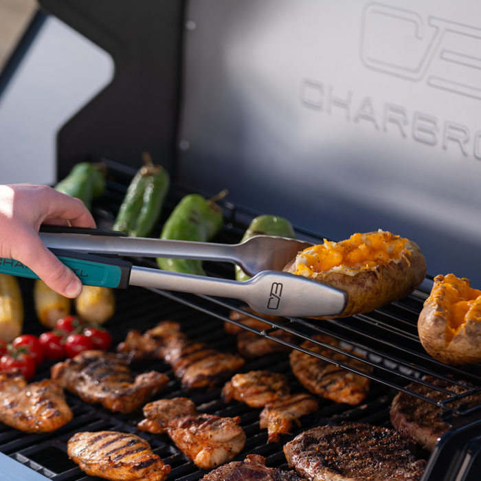 Silver Char-Broil tongs holding a grilled potato with cheese, near a grill with vegetables and meat.