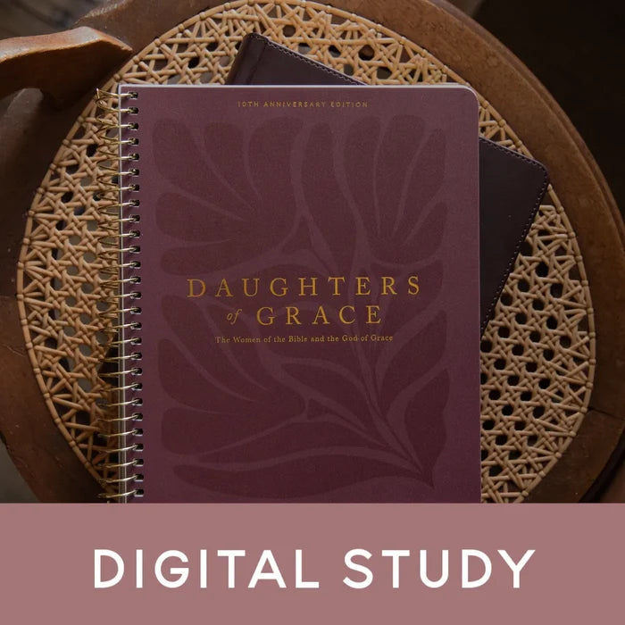 A spiral-bound book titled "Daughters of Grace" in a deep burgundy cover with gold lettering, placed on a woven round tray. A dark bookmark or card is tucked behind it.