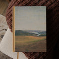 A hardcover bible with a landscape painting cover, resting on a cozy brown knitted blanket over a light-colored cushion.