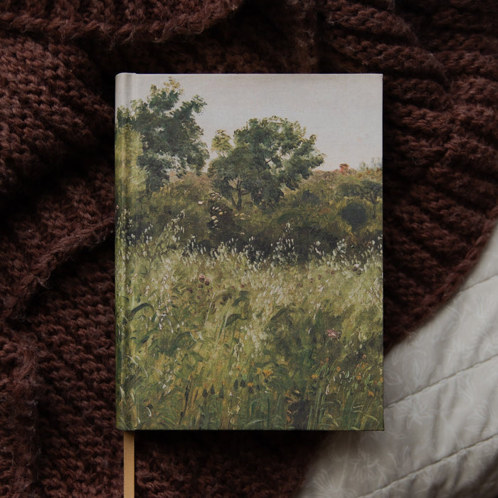 A hardcover notebook featuring an impressionist landscape painting of a green grassy field and trees, styled on a dark brown chunky knit blanket next to a white quilted pillow.