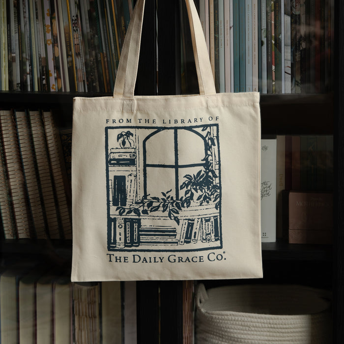 An off-white canvas tote bag with a dark blue graphic print of a library scene and the text 'FROM THE LIBRARY OF THE DAILY GRACE CO.' hanging in front of a bookshelf.