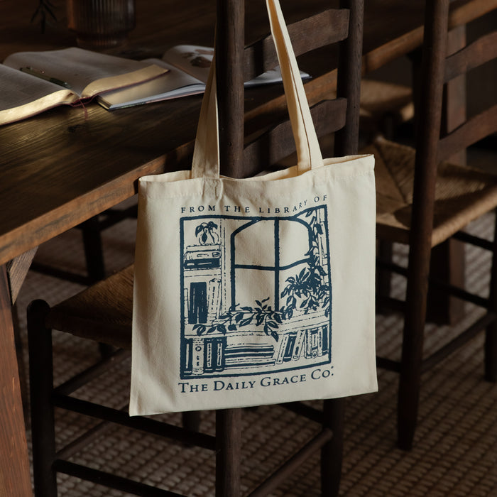 A cream canvas tote bag with a dark blue graphic print hanging from a wooden chair in a cozy, dimly lit library or study setting. The graphic features an illustration of a bookshelf and window with the text 'FROM THE LIBRARY OF THE Daily Grace Co.