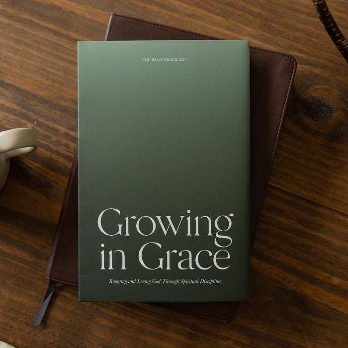 A hardcover book titled 'Growing in Grace' by Dr. Mark Deas lies on a dark brown leather journal, styled on a rustic wooden table next to a white ceramic coffee mug.