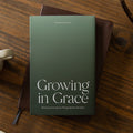 A hardcover book titled 'Growing in Grace' by Dr. Mark Deas lies on a dark brown leather journal, styled on a rustic wooden table next to a white ceramic coffee mug.
