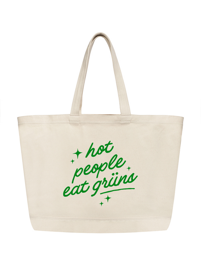 A natural canvas tote bag with long handles features green cursive text reading "hot people eat grüns" surrounded by small star accents, shown on a white background.
