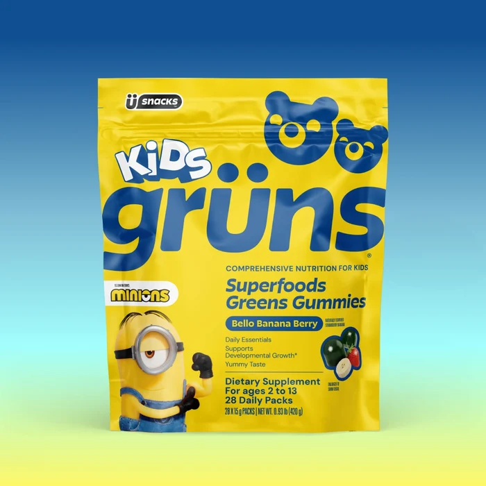 A yellow bag of Kids Grüns Superfoods Greens Gummies in "Bello Banana Berry" flavor features a Minions character on the front, labeled as a dietary supplement for ages 2 to 13 with 28 daily packs, set against a blue-to-yellow gradient background.