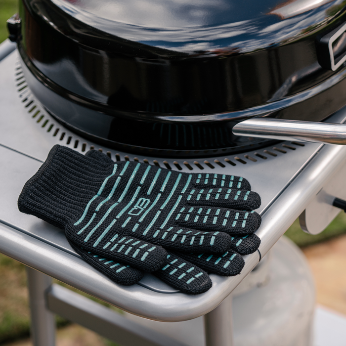Black heat-resistant gloves with teal accents on a grill handle.