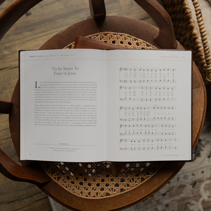 The I need Thee Every Hour book opened to the Tis so Sweet to trust in Jesus page on a wicker chair