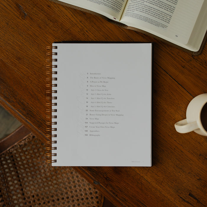 Identity in Christ Verse Map Journal opened to the table of contents page on a wooden table near a coffee mug and an open bible