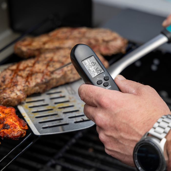 Black digital meat thermometer displaying temperature on a grill with steaks.