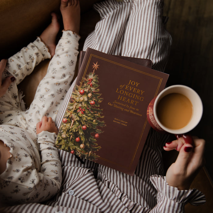 A cozy lifestyle scene showing a person in striped pajamas holding a brown hardcover book titled 'Joy of Every Longing Heart' with a Christmas tree illustration, while a baby in floral pajamas reaches toward the book and a hand holds a mug of tea.