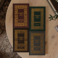 Four hardcover books titled 'Knowing God' arranged on a round wooden table, with two in dark brown and two in dark green, each featuring ornate gold decorative borders and gold lettering. The books are styled in a cozy home environment with a patterned rug, a plant, and other books visible.