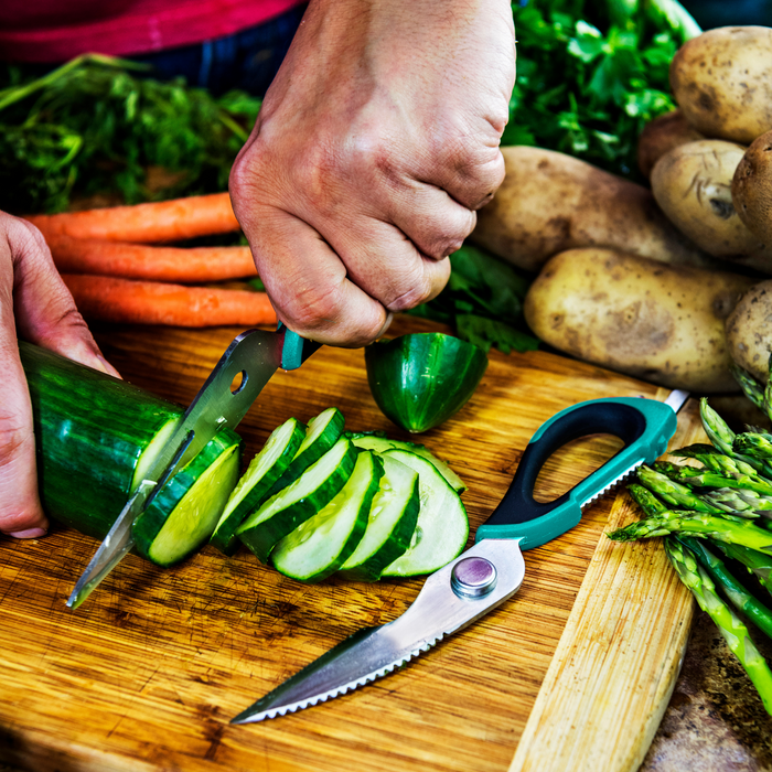 Green-handled kitchen shears cutting cucumber on wooden cutting board.
