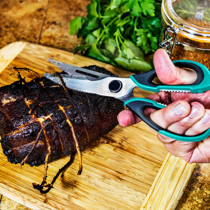 Green-handled kitchen shears cutting grilled meat on wooden board.