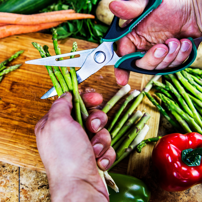 Green-handled kitchen shears cutting asparagus on wooden board.