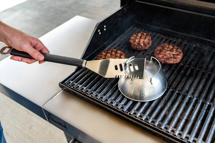 Silver and black multi-tool grill spatula with burger patties on a black grill.