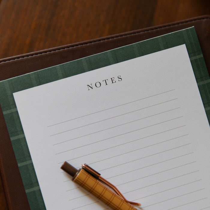 A close-up of a lined notes pad with a green plaid border, placed on a brown leather folio with a tan plaid pen resting on it, set on a wooden surface.