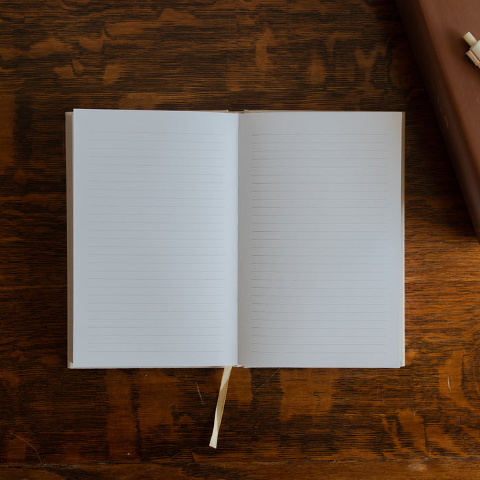 The Notes Lined Journal opened to a blank page on a wooden table next to a bible with a white pen on it