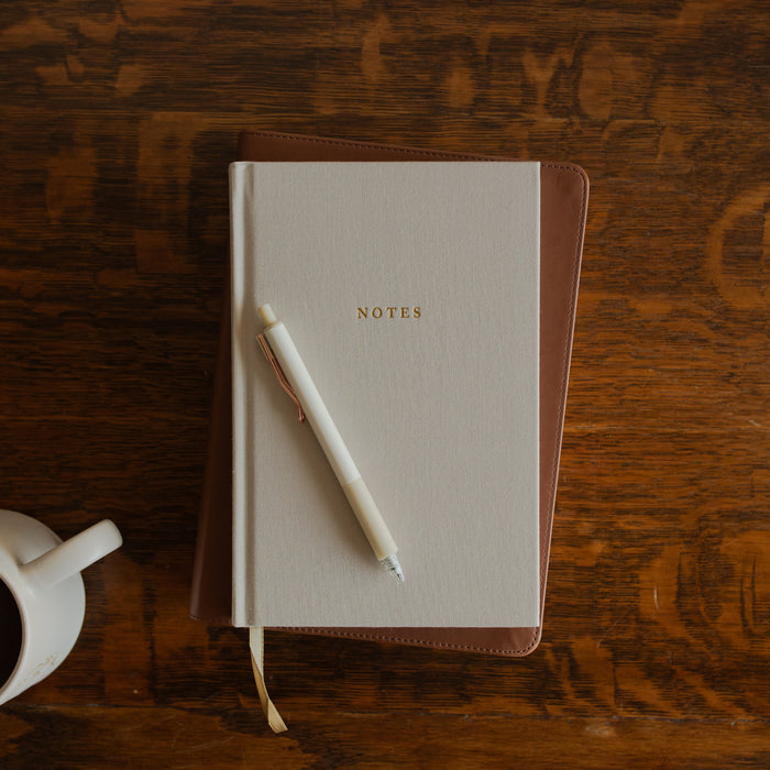 The Notes Lined Journal cover on a wooden table on a bible with a white pen on it near a white coffee cup