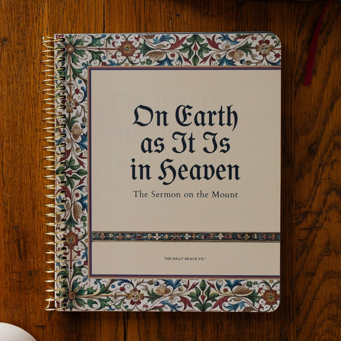 Notebook titled 'On Earth as It Is in Heaven' on a wooden surface with an open book and coffee cup.