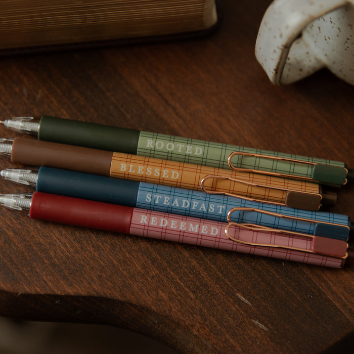 Four plaid retractable pens in green, brown, blue, and pink, stacked diagonally on a dark wooden table. Each pen has a different color body with a gold-tone pocket clip and is printed with a religious phrase: 'ROOTED', 'BLESSED', 'STEADFAST', and 'REDEEMED'.