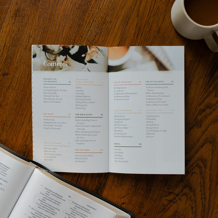 The Prayers for your Day opened to the table of contents page on a wooden table near a coffee cup