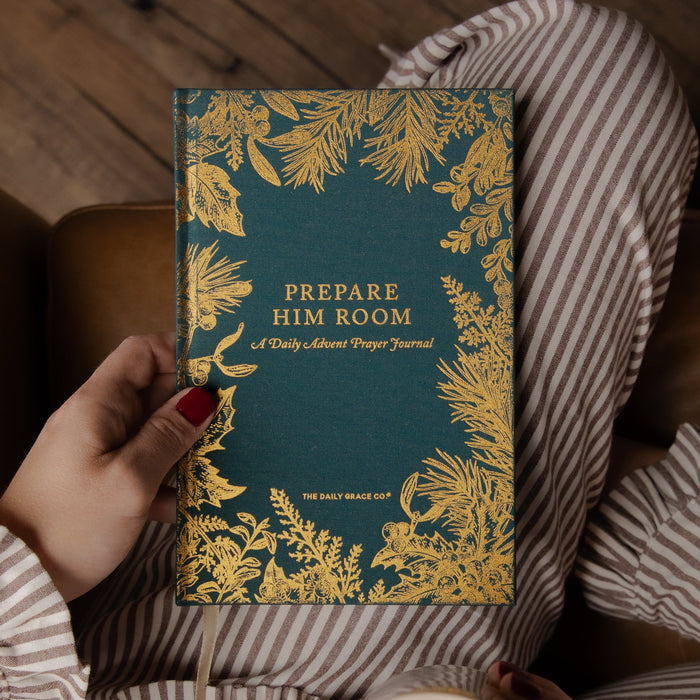 A person in striped pajamas holds a teal journal with ornate gold botanical borders and the title 'PREPARE HIM ROOM' on the cover, photographed in a cozy indoor setting with a wooden floor and chair visible.