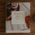 A spiral-bound notebook titled 'Search the Word' lies on a wooden table, surrounded by an open Bible, a ceramic mug, and part of another book. The notebook cover features a warm-toned photograph with a cream title panel.