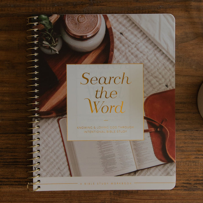 A spiral-bound notebook titled 'Search the Word' lies on a wooden table, surrounded by an open Bible, a ceramic mug, and part of another book. The notebook cover features a warm-toned photograph with a cream title panel.