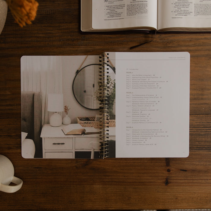 An open spiral-bound journal or planner lies on a wooden table, showing a bedroom photo on the left page and a list of verses from Psalm 34 on the right page. An open book and part of a mug are visible around it in a styled flat-lay setting.