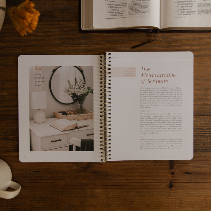 An open spiral-bound book or journal lies on a wooden tabletop, showing a page titled 'The Metanarrative of Scripture' with text and a left-page photo of a styled interior scene. An open book and a small vase are partially visible around it.