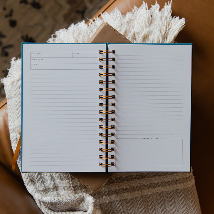 An open spiral-bound planner or journal with lined pages, photographed from above on a brown leather surface with a cream fringed textile and knit blanket underneath.